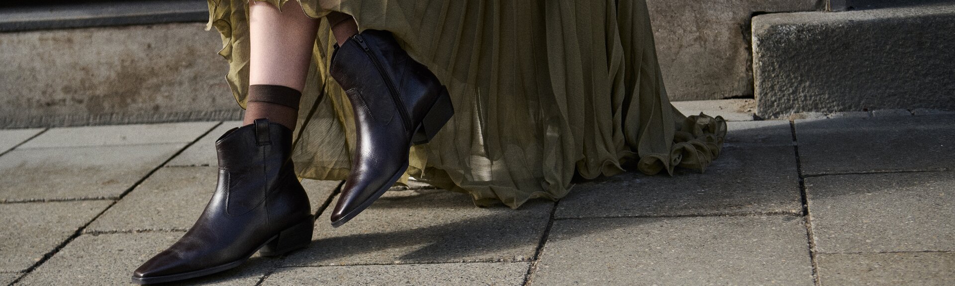 Een vrouw draagt donkerbruine dames-enkellaarsjes met spitse neus en lage hak bij een lange, zwierige olijfgroene rok; ze staat op een geplaveide stoep voor stenen treden. | © Gabor Shoes GmbH, Duitsland
