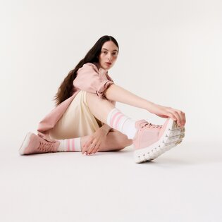 Une femme est assise par terre dans une pièce lumineuse et porte des baskets roses, des chaussettes rayées, une jupe crème et une veste rose. | © Gabor Shoes GmbH, Allemagne