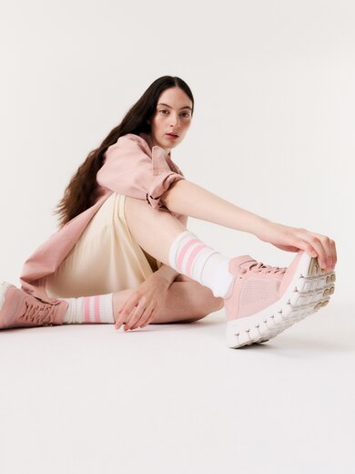 Une femme est assise par terre dans une pièce lumineuse et porte des baskets roses, des chaussettes rayées, une jupe crème et une veste rose. | © Gabor Shoes GmbH, Allemagne