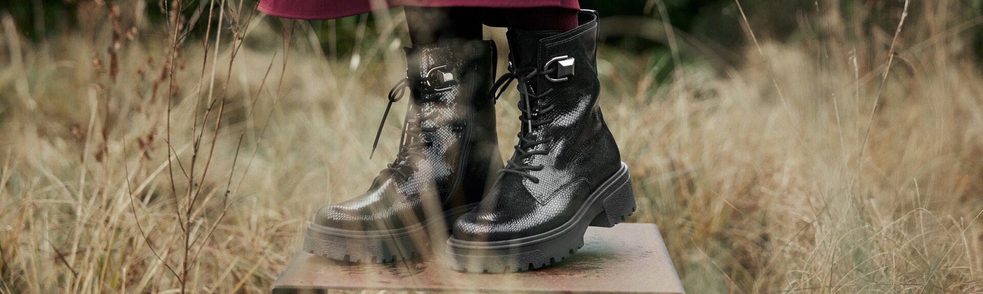 Des bottines noires texturées à semelle épaisse sont posées sur une plaque en métal dans de l’herbe sèche et haute. La femme porte un pantalon bordeaux. | © Gabor Shoes GmbH, Allemagne