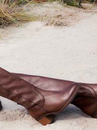 Une femme est assise détendue dans des vêtements bruns et des bottes brunes sur du sable, avec de l’herbe et des plantes vertes en arrière-plan. | © Gabor Shoes GmbH, Allemagne