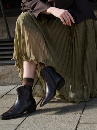 Een vrouw draagt donkerbruine dames-enkellaarsjes met spitse neus en lage hak bij een lange, zwierige olijfgroene rok; ze staat op een geplaveide stoep voor stenen treden. | © Gabor Shoes GmbH, Duitsland