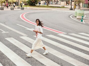 Une femme traverse une large rue sur un passage piéton. Elle porte un t-shirt blanc, une longue jupe claire et des baskets blanches, et tient un smartphone dans la main. À l’arrière-plan, on aperçoit des arbres et quelques piétons. | © Gabor Shoes GmbH, Allemagne