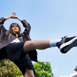 Une femme se tient à l’extérieur, porte une veste foncée, une jupe noire et des baskets noires, une jambe est levée en l’air. | © Gabor Shoes GmbH, Allemagne