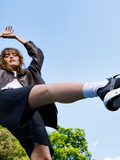 Une femme se tient à l’extérieur, porte une veste foncée, une jupe noire et des baskets noires, une jambe est levée en l’air. | © Gabor Shoes GmbH, Allemagne