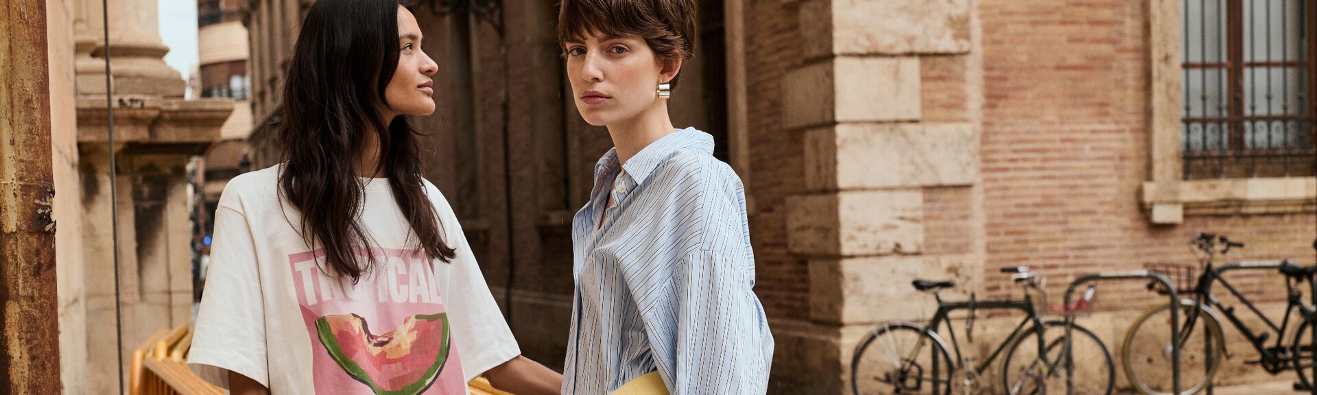 Deux femmes se tiennent côte à côte devant un vieux bâtiment ; l’une porte un t-shirt blanc avec un imprimé pastèque et l’inscription « Tropical », l’autre une chemise bleu clair. À l’arrière-plan, des vélos sont appuyés contre une barrière. | © Gabor Shoes GmbH, Allemagne