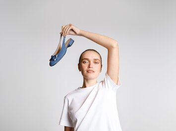 Une femme en t-shirt blanc tient une chaussure bleue avec petit talon et nœud en l’air, devant un fond clair. | © Gabor Shoes GmbH, Allemagne