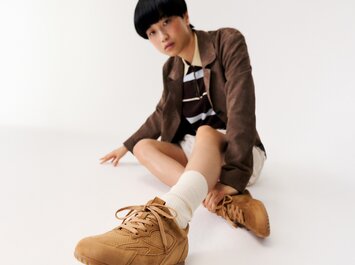 Une femme est assise par terre dans une pièce blanche et porte des baskets marron, des chaussettes blanches, une jupe beige, un haut rayé et un blazer marron. | © Gabor Shoes GmbH, Allemagne