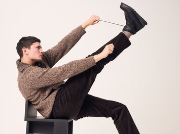 Un homme est assis sur une chaise, lève une jambe et noue une bottine noire à lacets. La personne porte un pull marron et un pantalon sombre. L’arrière-plan est clair et neutre. | © Gabor Shoes GmbH, Allemagne