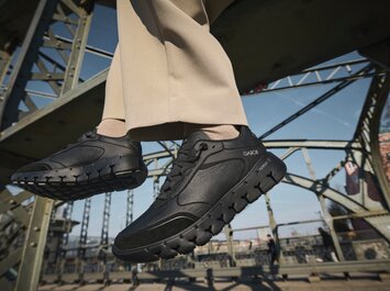 En man sitter på en stålbro, bär ljusbeige byxor och svarta Gabor-sneakers, med fötterna hängande över gatan. | © Gabor Shoes GmbH, Tyskland