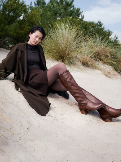 Une femme est assise détendue dans une tenue élégante brune et des bottes brunes sur un sol sableux entre dunes et herbes. En arrière-plan, on voit des buissons verts et des arbres. | © Gabor Shoes GmbH, Allemagne