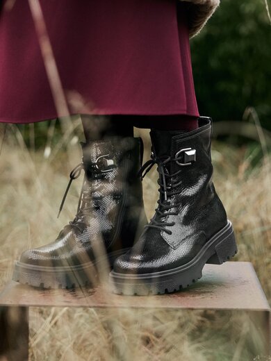 Des bottines noires texturées à semelle épaisse sont posées sur une plaque en métal dans de l’herbe sèche et haute. La femme porte un pantalon bordeaux. | © Gabor Shoes GmbH, Allemagne