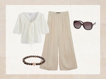 On voit une blouse blanche avec des liens à nouer, un pantalon beige à la coupe ample, des lunettes de soleil marron foncé et un bracelet en perles marron foncé. | © aboutyou.de 