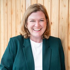 Personne aux cheveux blonds mi-longs en blazer vert et t-shirt blanc devant un mur en bois. | © Gabor Shoes GmbH, Allemagne