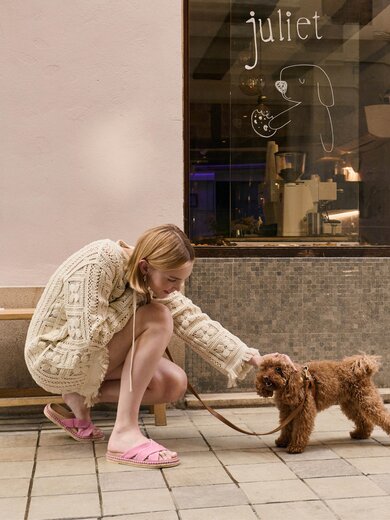 Une femme en robe en tricot beige et pantoufles roses s’agenouille sur un trottoir et caresse un petit chien brun en laisse. À l’arrière-plan, il y a un banc et une vitrine portant l’inscription « juliet » ainsi qu’un dessin de chien. | © Gabor Shoes GmbH, Allemagne