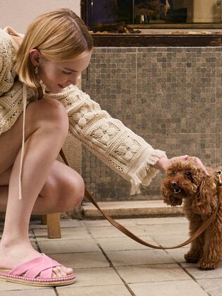 Une femme en robe en tricot beige et pantoufles roses s’agenouille sur le trottoir et caresse un petit chien brun au pelage bouclé. À côté se trouvent un banc en bois avec un gobelet dessus et un vélo couleur menthe. | © Gabor Shoes GmbH, Allemagne