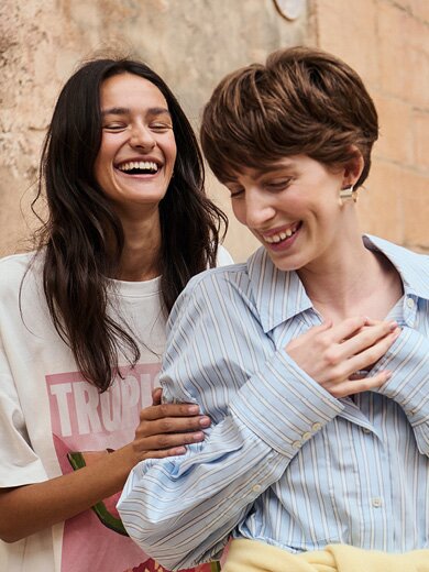 myGabor | Zwei lachende Frauen stehen leicht versetzt hintereinander vor einer alten Steinmauer, Nahaufnahme der Oberkörper und Gesichter | © Gabor Shoes GmbH, Rosenheim